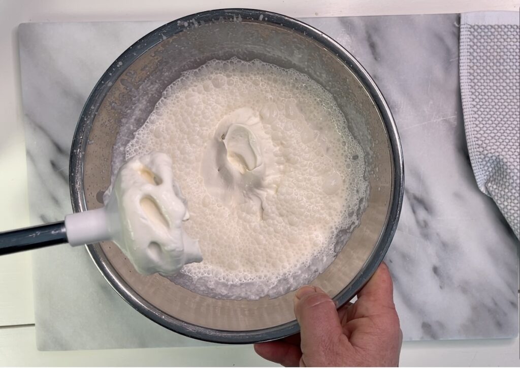 Hand holding a bowl of whipped cream next to a whisk attachment on a marble surface with a textured cloth.