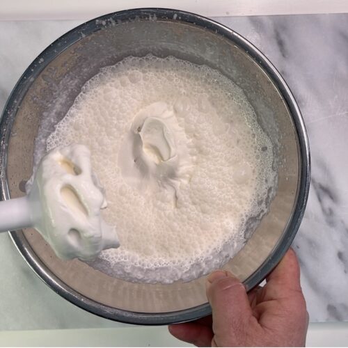 Hand holding a bowl of whipped cream next to a whisk attachment on a marble surface with a textured cloth.