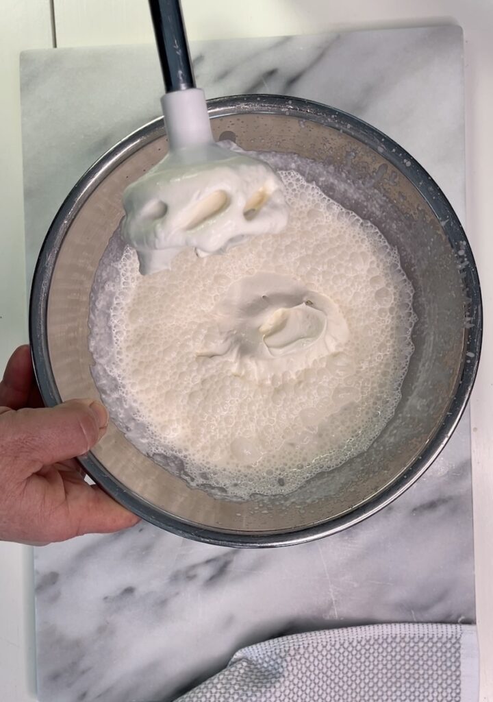 A hand holds a mixing bowl with whipping cream being mixed. A spatula lifts some of the cream, displaying its texture. The bowl is on a marble surface.
