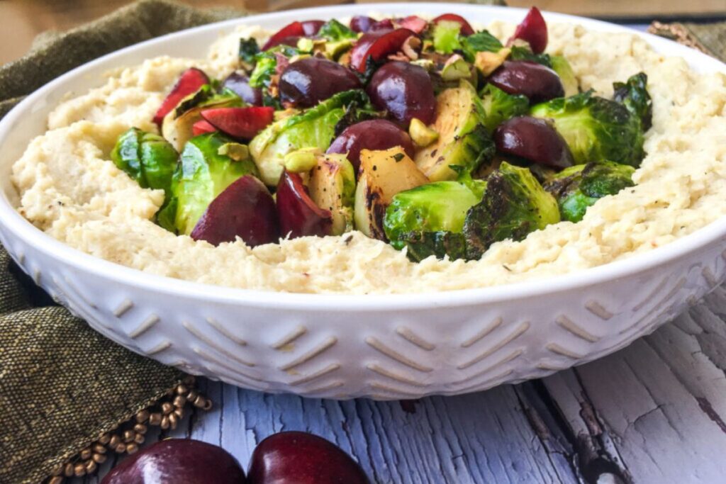 A white bowl of hummus topped with roasted Brussels sprouts, cherries, and nuts. It sits on a wooden surface with a green cloth.