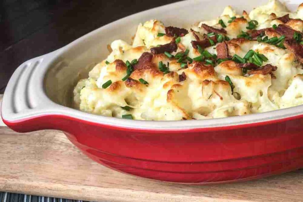 A red baking dish filled with a baked cauliflower casserole topped with crispy bacon and chopped chives.