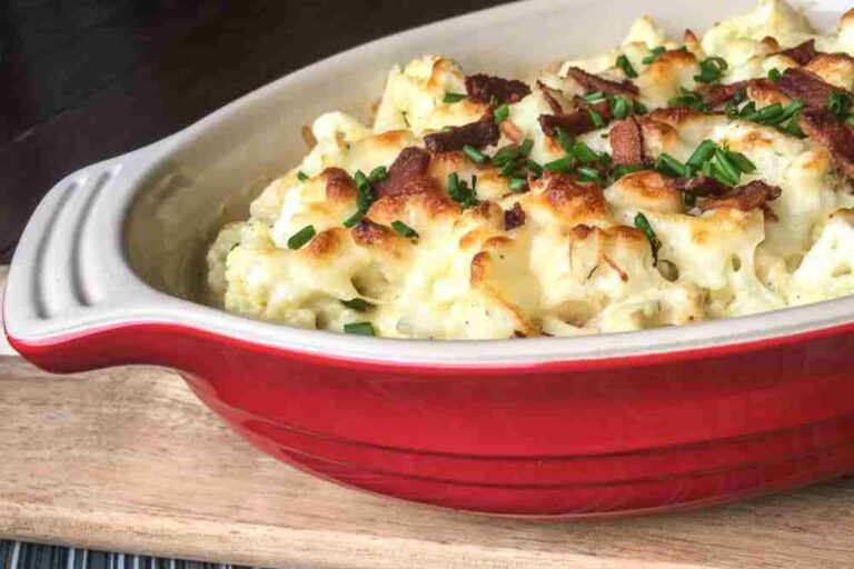 A red baking dish filled with a baked cauliflower casserole topped with crispy bacon and chopped chives.