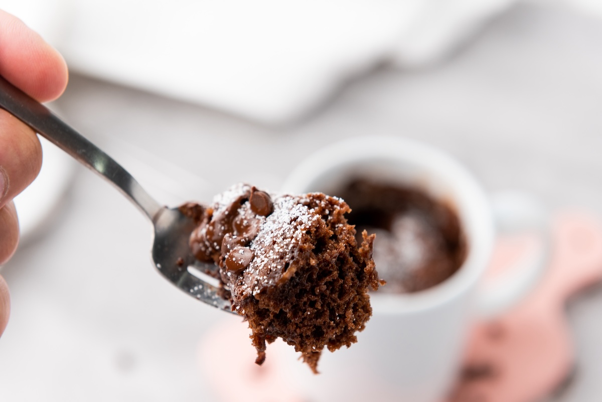 A fork holds a bite of chocolate cake topped with powdered sugar, with a mug cake in the background.