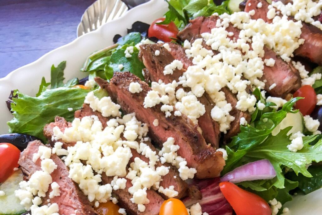 A plate of sliced steak on a bed of mixed greens, cherry tomatoes, cucumber, red onion, and crumbled cheese.