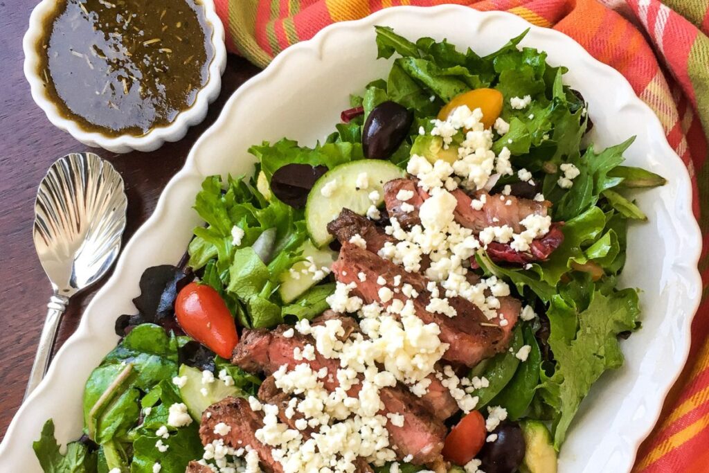 Salad with sliced steak, cherry tomatoes, cucumbers, and crumbled cheese on a white platter. A small dish of dressing is beside it, with a spoon and a multicolored napkin nearby.