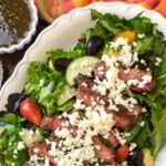 Salad with sliced steak, cherry tomatoes, cucumbers, and crumbled cheese on a white platter. A small dish of dressing is beside it, with a spoon and a multicolored napkin nearby.