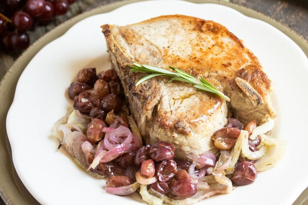 A cooked pork chop resting on a bed of sautéed onions and grapes, garnished with a sprig of rosemary, served on a white plate.