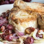 A cooked pork chop on a white plate, served with sautéed onions and grapes.