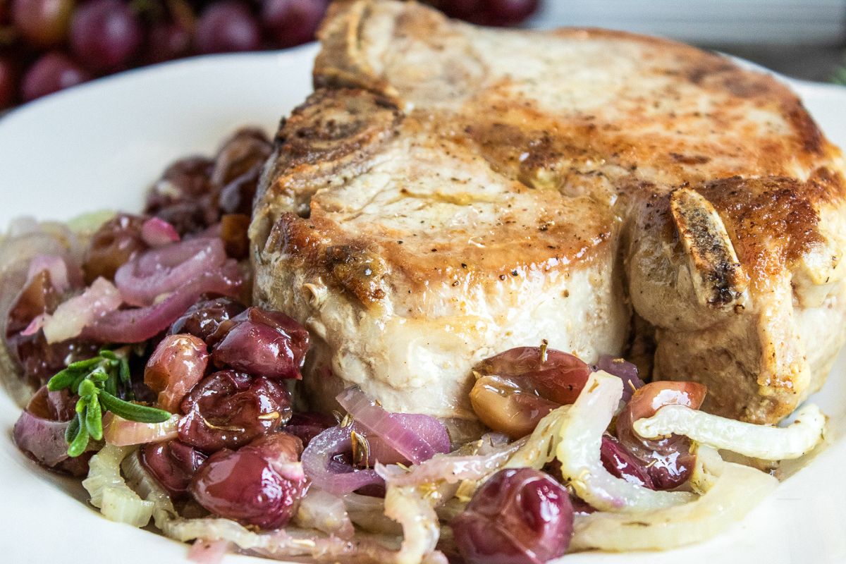 A cooked pork chop on a white plate, served with sautéed onions and grapes.