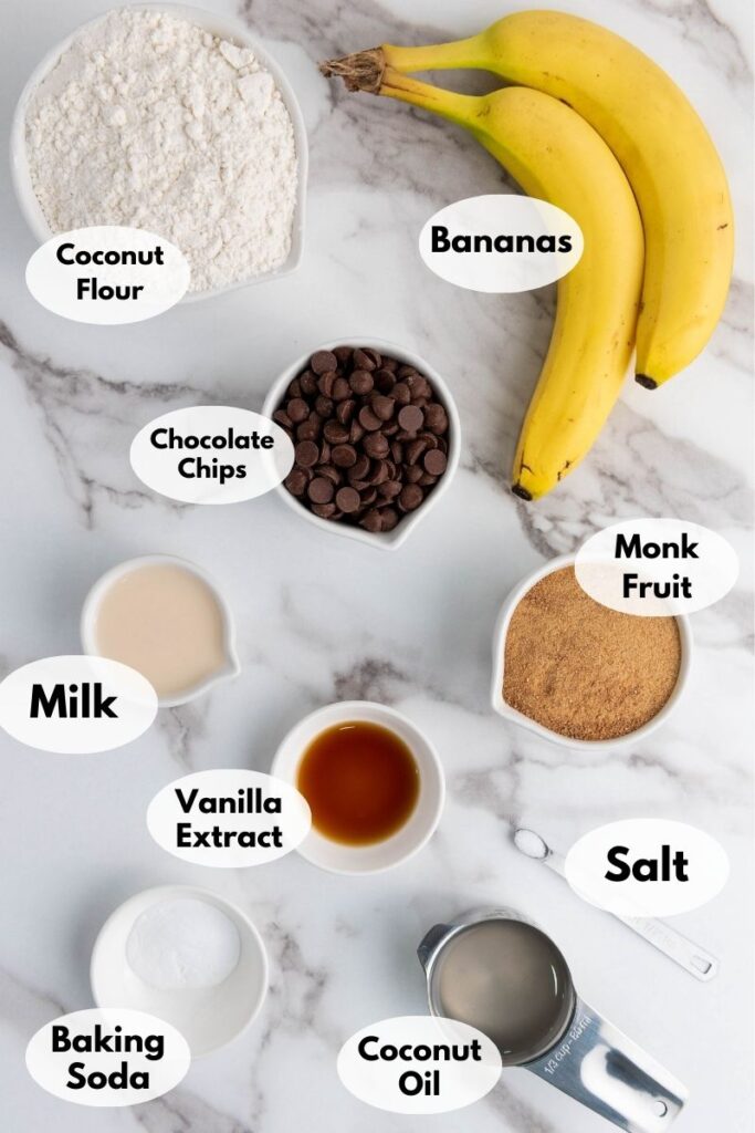 Overhead view of ingredients arranged on a marble surface: coconut flour, bananas, chocolate chips, monk fruit, milk, vanilla extract, baking soda, coconut oil, and salt.