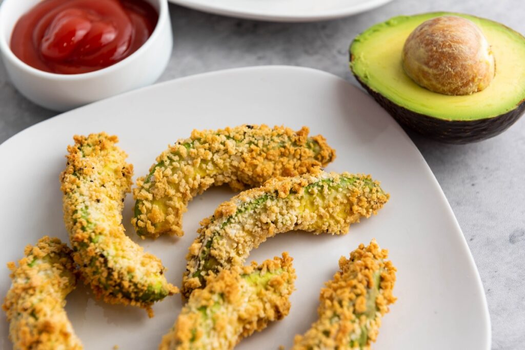 Crispy avocado fries on a white plate with a halved avocado and a small bowl of ketchup in the background.