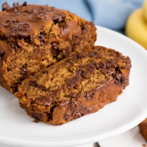 A sliced loaf of chocolate chip banana bread is displayed on a white plate, with bananas in the background.