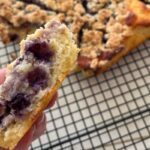 A close-up of a hand holding a slice of blueberry cake with a crumb topping. The whole cake is on a cooling rack in the background.