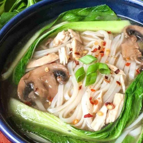 Bowl of soup with rice noodles, bok choy, mushroom slices, tofu pieces, chili flakes, and chopped green onions.