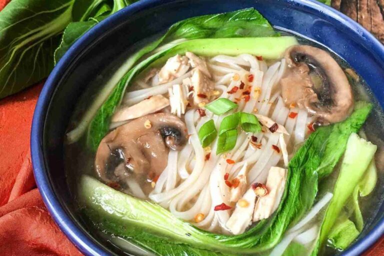 Bowl of soup with rice noodles, bok choy, mushroom slices, tofu pieces, chili flakes, and chopped green onions.