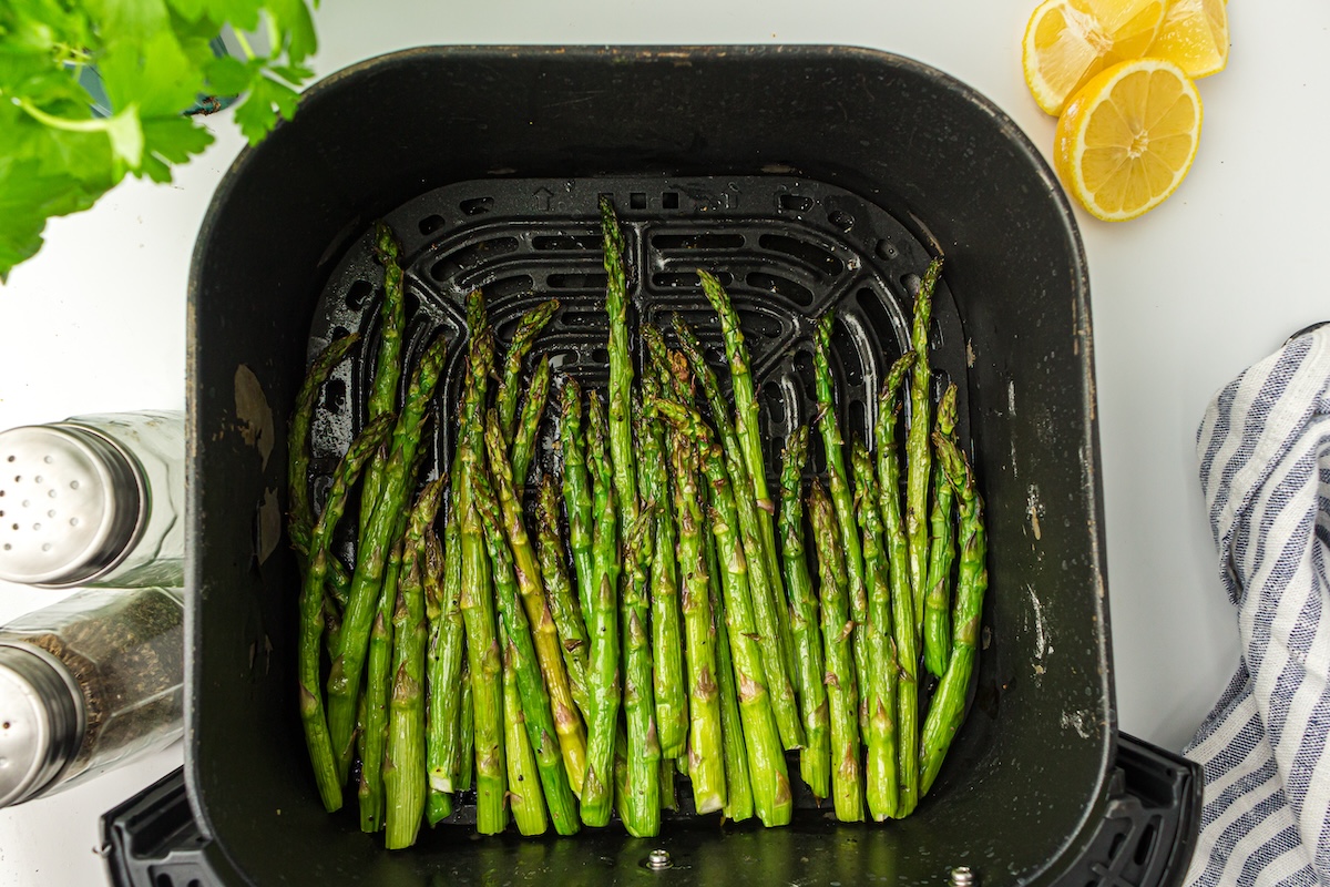 Asparagus spears arranged in an air fryer basket with a striped cloth, sliced lemons, and salt and pepper shakers nearby on a white surface.