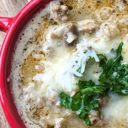 A red bowl filled with creamy turkey mushroom soup, topped with chopped greens and grated cheese, rests on a wooden surface.