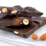 Pieces of dark chocolate bark with whole almonds, stacked on white parchment paper, with a few loose almonds beside them.