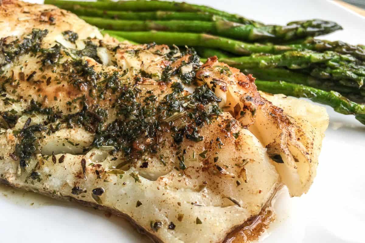 Grilled fish fillet with herbs served alongside cooked asparagus on a white plate.