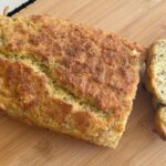 A loaf of homemade bread on a wooden cutting board with one thick slice cut off, showing a textured, seeded interior.