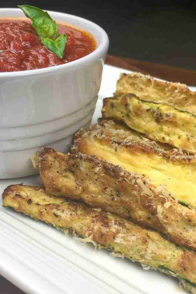 Zucchini fries coated in breadcrumbs and grated cheese, served on a white plate with a side of marinara sauce topped with fresh basil in a small white bowl.