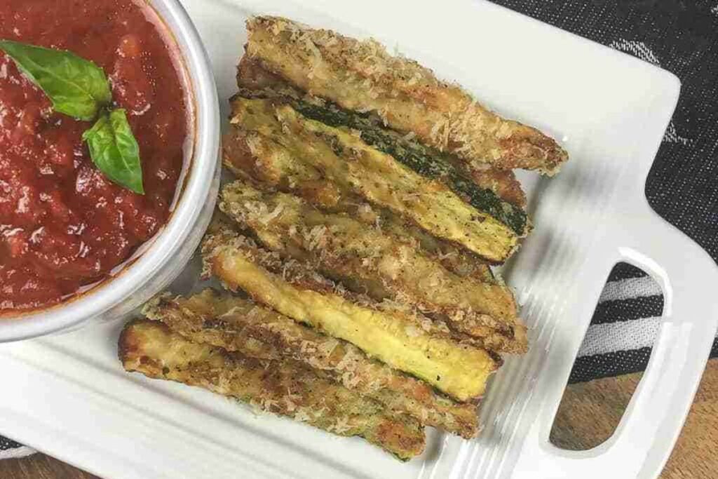Plate of breaded zucchini fries next to a bowl of marinara sauce with two basil leaves, served on a white rectangular dish.