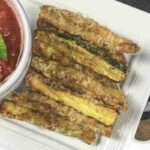 Plate of breaded zucchini fries next to a bowl of marinara sauce with two basil leaves, served on a white rectangular dish.