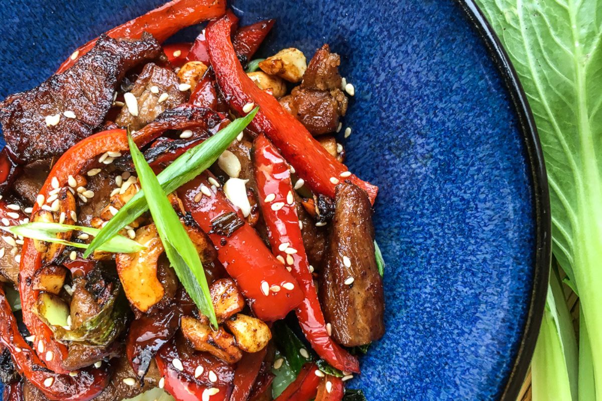 Stir-fried beef with red bell peppers, zucchini, and sesame seeds, reminiscent of a pork cashew stir fry, garnished with green onions on a blue plate.