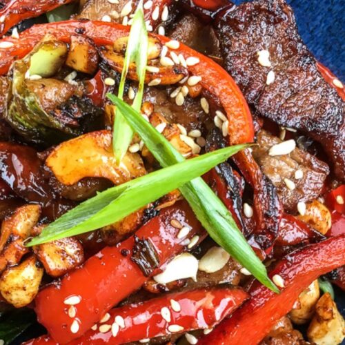 Stir-fried beef with red peppers, sliced garlic, scallions, sesame seeds, and zucchini on a dark blue plate, offering a delightful twist akin to a pork cashew stir fry.
