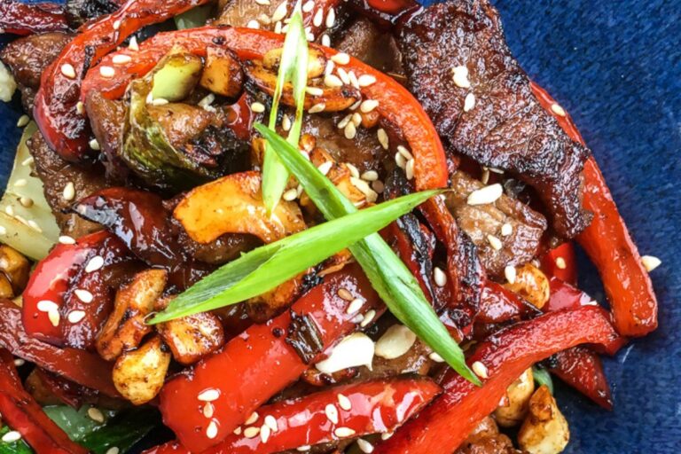 Stir-fried beef with red peppers, sliced garlic, scallions, sesame seeds, and zucchini on a dark blue plate, offering a delightful twist akin to a pork cashew stir fry.