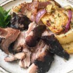 Plate with sliced roasted meat, roasted potatoes with red onions, and a sprig of fresh parsley, served on a white dish.