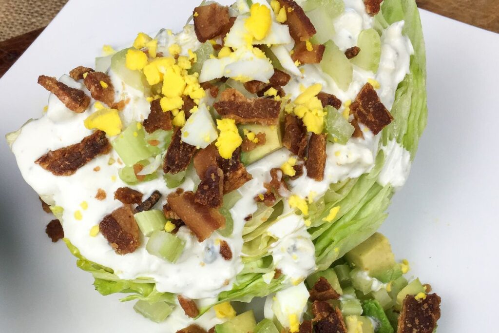 Wedge salad with iceberg lettuce, chopped bacon, crumbled hard-boiled egg, diced celery, and avocado, topped with creamy dressing, on a white plate.