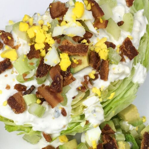 Wedge salad with iceberg lettuce, chopped bacon, crumbled hard-boiled egg, diced celery, and avocado, topped with creamy dressing, on a white plate.