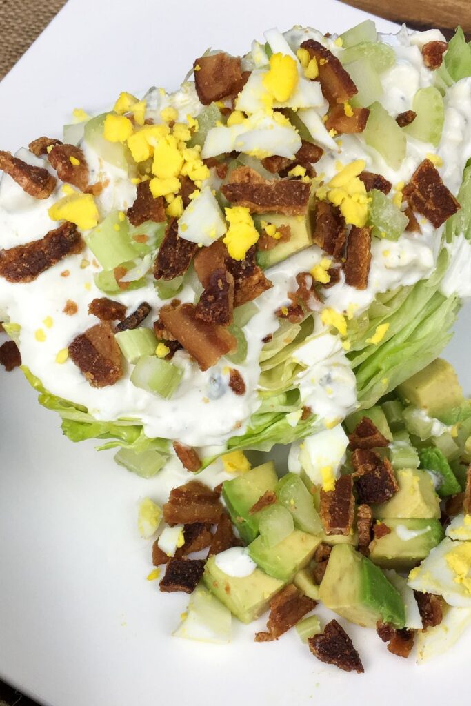 Wedge salad topped with blue cheese dressing, bacon bits, chopped boiled egg, celery, and diced avocado, served on a white plate.