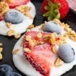 Yogurt rounds topped with sliced strawberries, whole blueberries, and granola are arranged on a black plate.