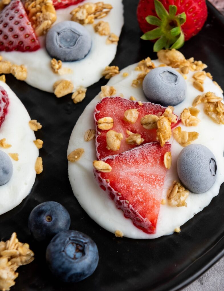Spoonfuls of yogurt topped with sliced strawberries, whole blueberries, and granola on a black plate.
