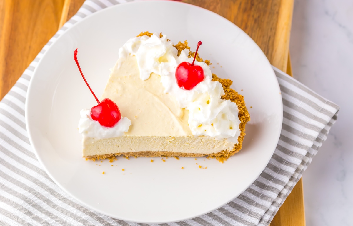 A slice of cream pie with a graham cracker crust, topped with whipped cream and two cherries, on a white plate placed on a striped napkin.