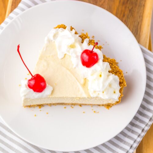 Slice of creamy dessert pie with whipped topping and two cherries on a white plate over a gray and white striped cloth.