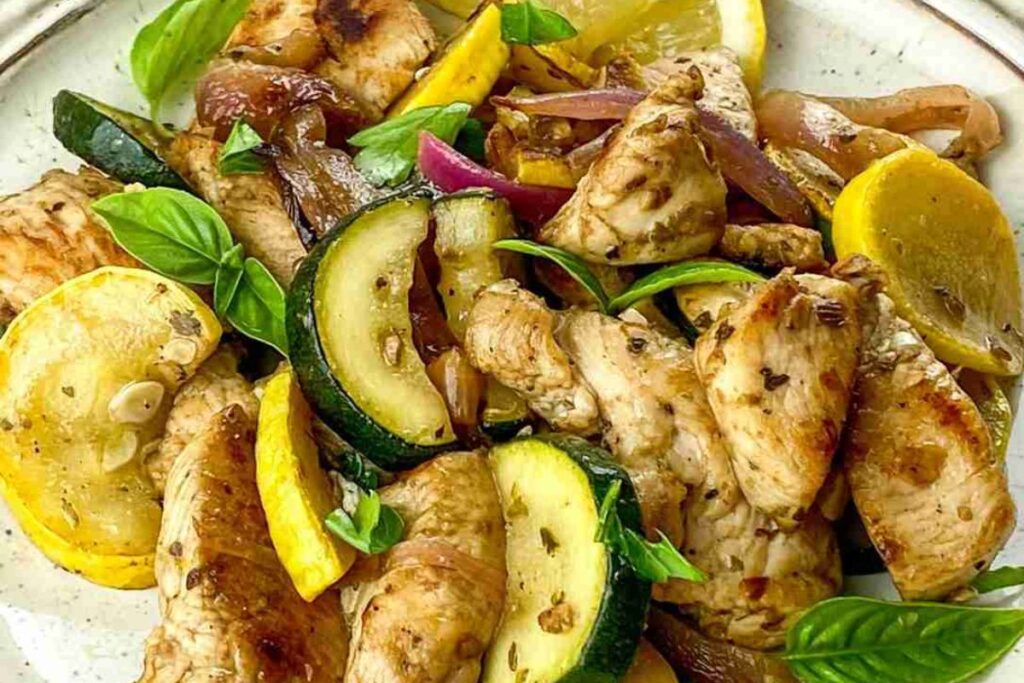 Grilled chicken pieces with sautéed zucchini, yellow squash, red onions, and fresh basil on a white plate.