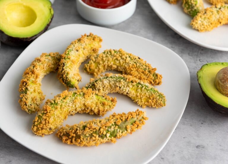 Six breaded avocado slices are arranged on a white plate, with halved avocados and a bowl of ketchup visible in the background.