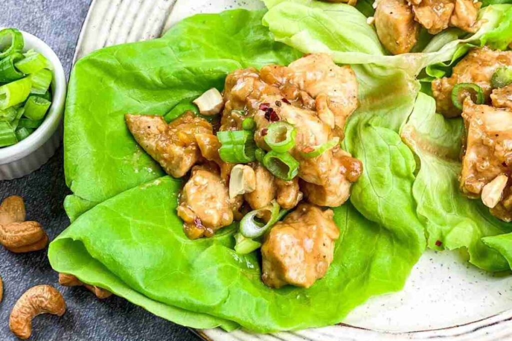 Chicken pieces with cashews and sliced green onions served on fresh lettuce leaves, with a small bowl of chopped green onions and some cashews on the side.