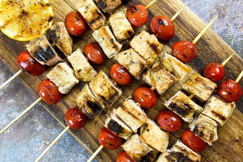 Grilled skewers with chunks of chicken and whole cherry tomatoes on a wooden board, with a grilled lemon half on the side.