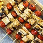 Grilled skewers with chunks of chicken and whole cherry tomatoes on a wooden board, with a grilled lemon half on the side.