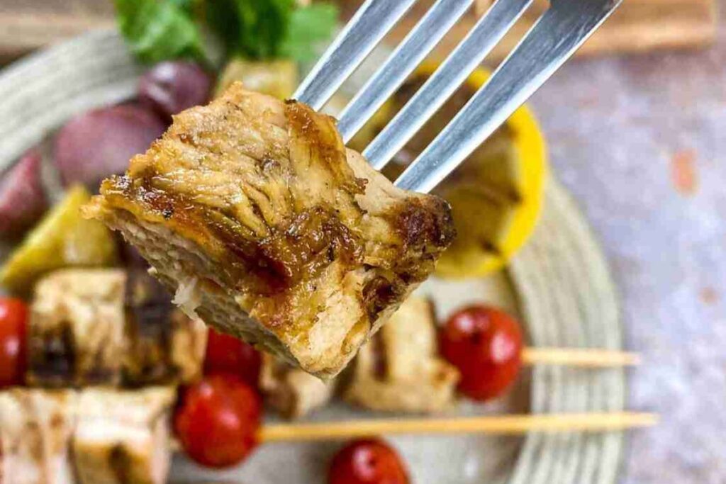 A close-up of a fork holding a grilled piece of chicken, with skewers of meat and cherry tomatoes visible in the blurred background.