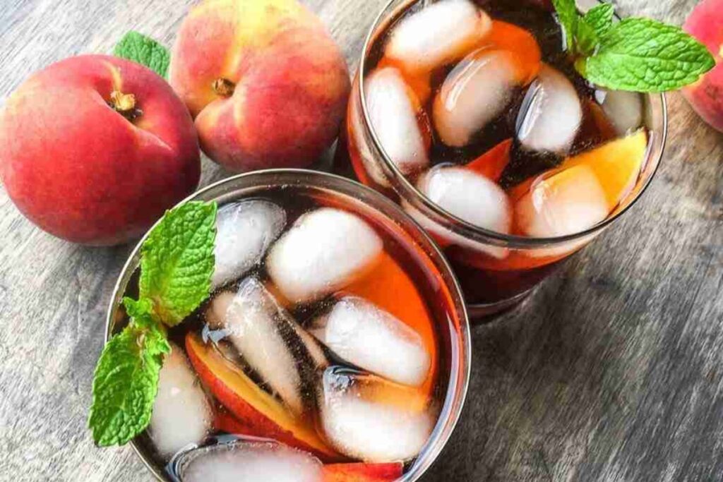 Two glasses of iced tea with peach slices and mint sprigs, next to whole peaches, on a wooden surface.