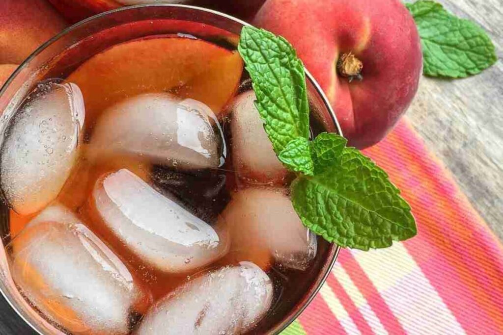 Glass of iced tea with ice cubes and a mint sprig, placed next to peaches on a colorful plaid cloth.