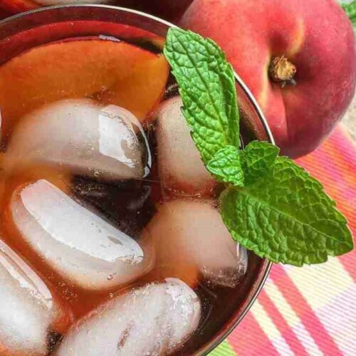 Glass of iced tea with ice cubes and a mint sprig, placed next to peaches on a colorful plaid cloth.