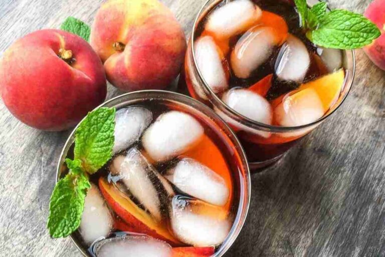 Two glasses of iced tea with peach slices and mint sprigs, next to whole peaches, on a wooden surface.