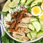 A salad bowl with sliced grilled chicken, hard-boiled eggs, cucumber, avocado, bacon, blue cheese, lime wedges, and lettuce greens, next to a striped cloth napkin.
