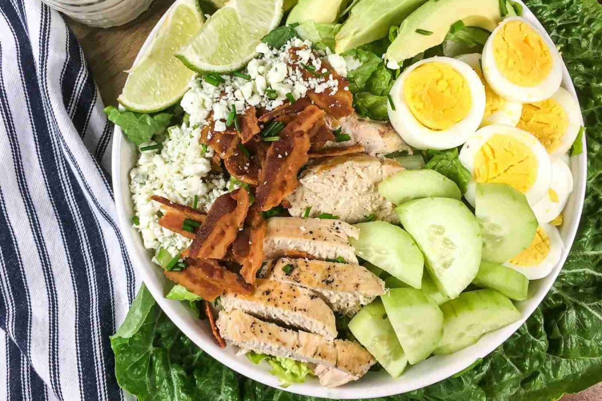 A bowl of salad with grilled chicken, bacon, hard-boiled eggs, avocado, cucumber, lettuce, cheese, lime wedges, and ranch dressing being spooned on top.